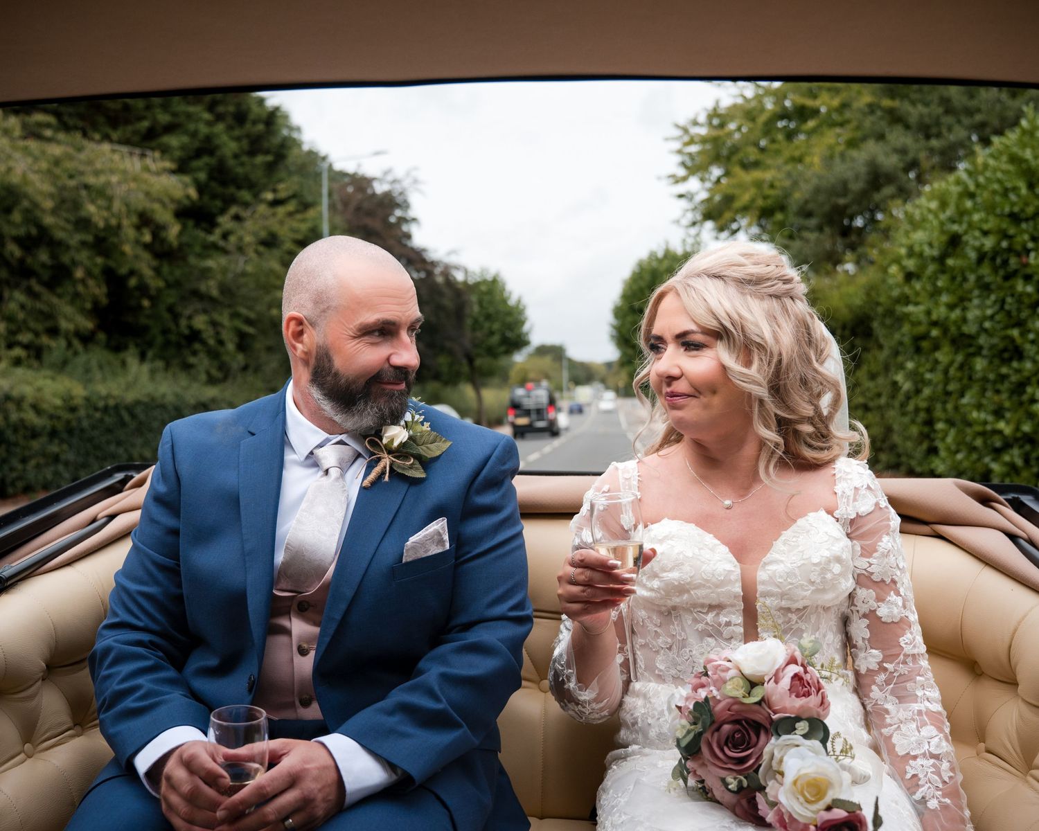 Bride and groom in wedding attire ride in an open-top car, holding champagne glasses, smiling on their wedding day.