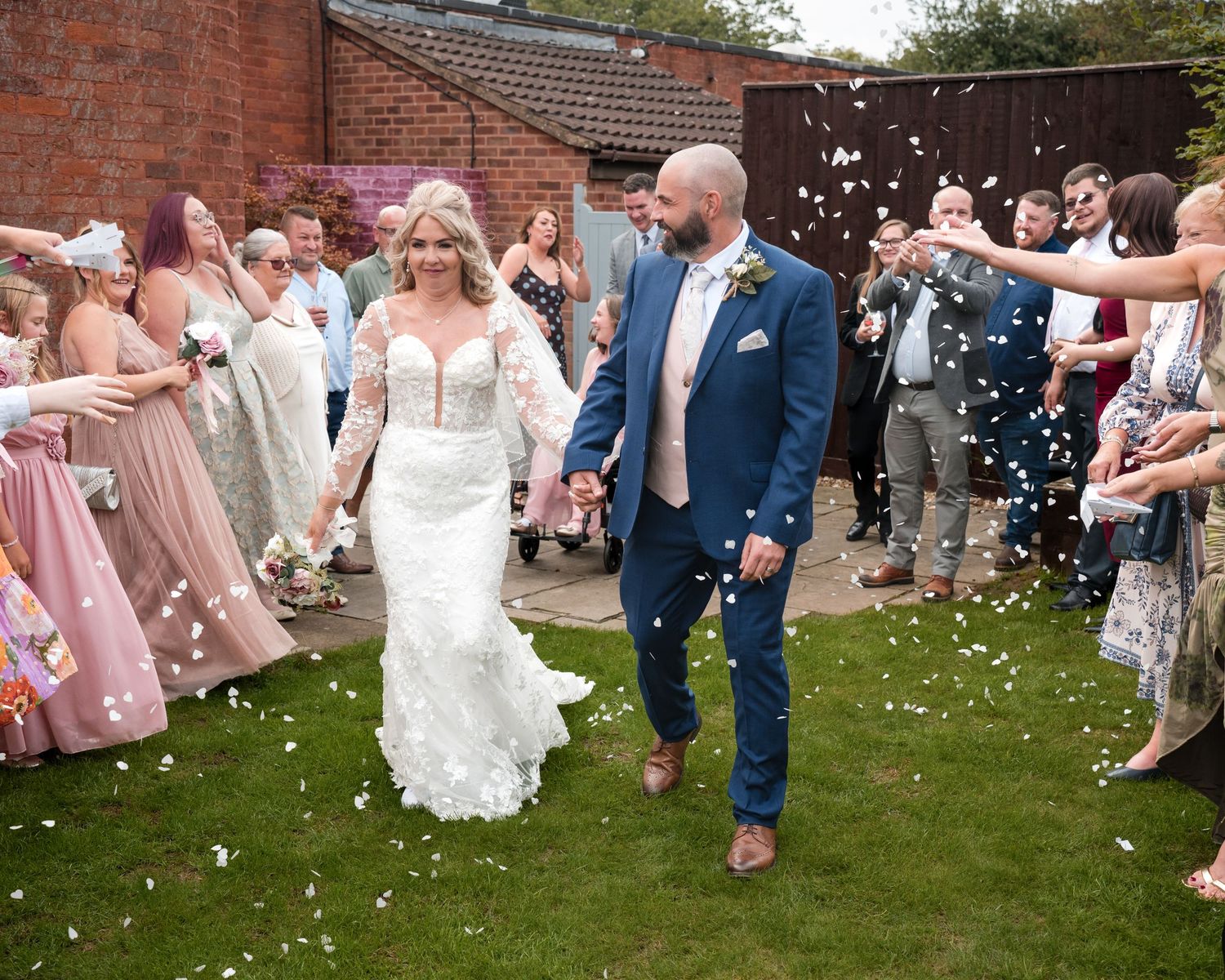 Bride and groom walk through confetti shower surrounded by smiling wedding guests outdoors on a sunny day.