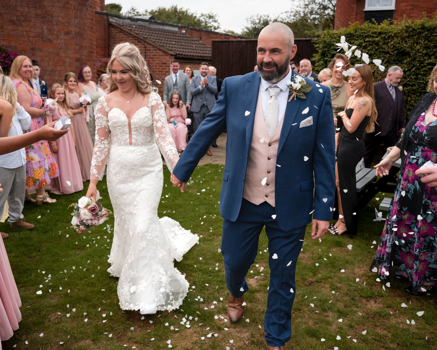 Bride and groom walk through confetti shower surrounded by cheering guests at outdoor wedding ceremony.