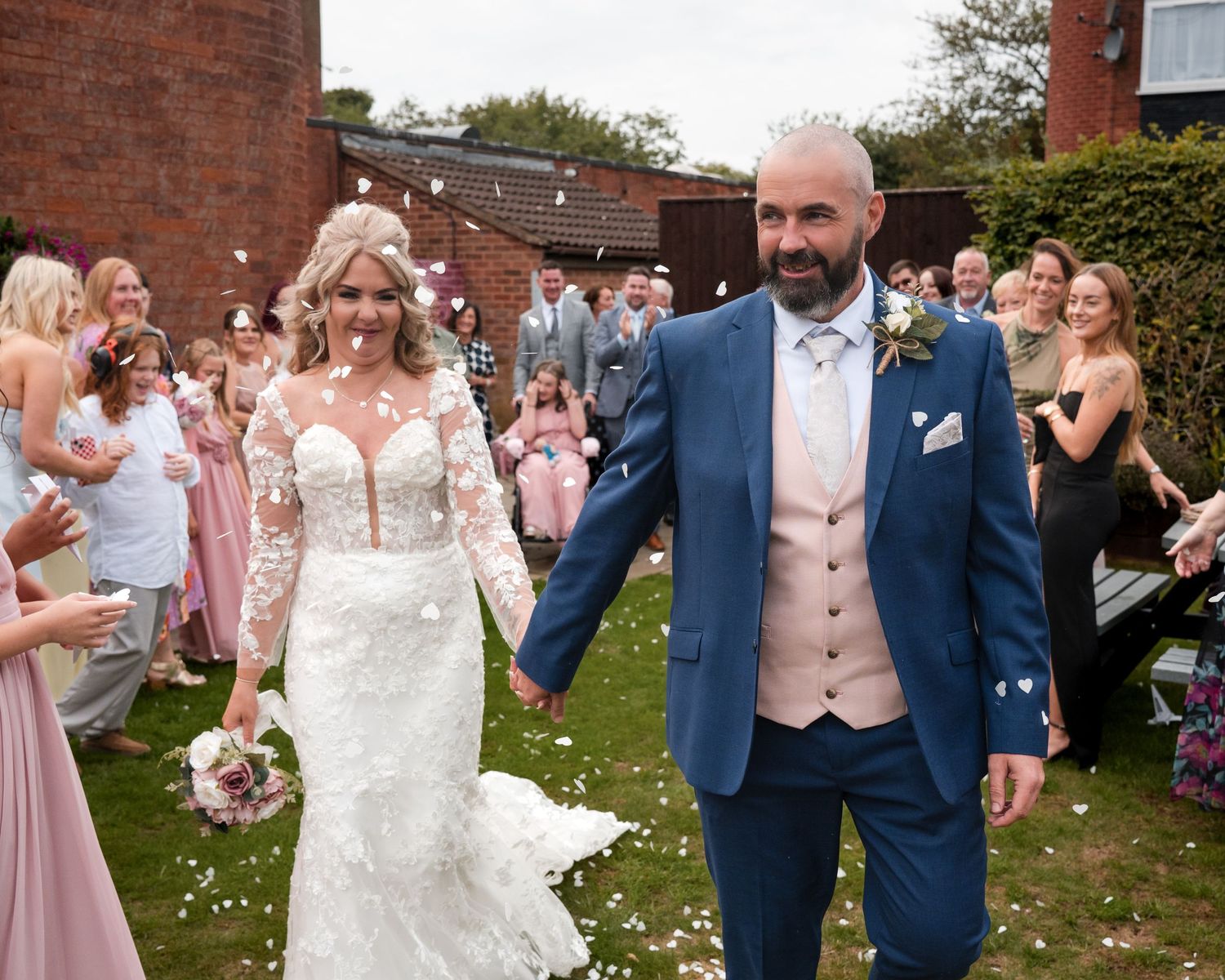 Bride and groom walk through confetti shower surrounded by cheering wedding guests outside a brick venue.