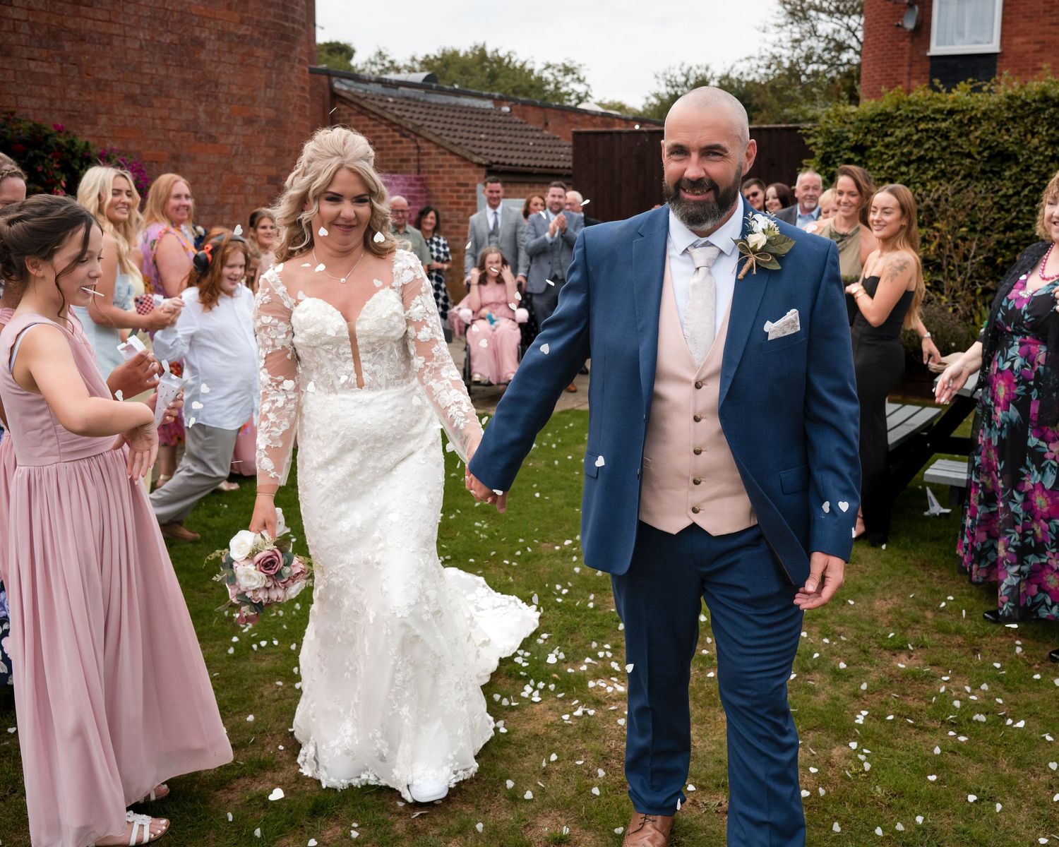 Bride and groom walk hand in hand through confetti shower surrounded by cheering wedding guests outdoors.