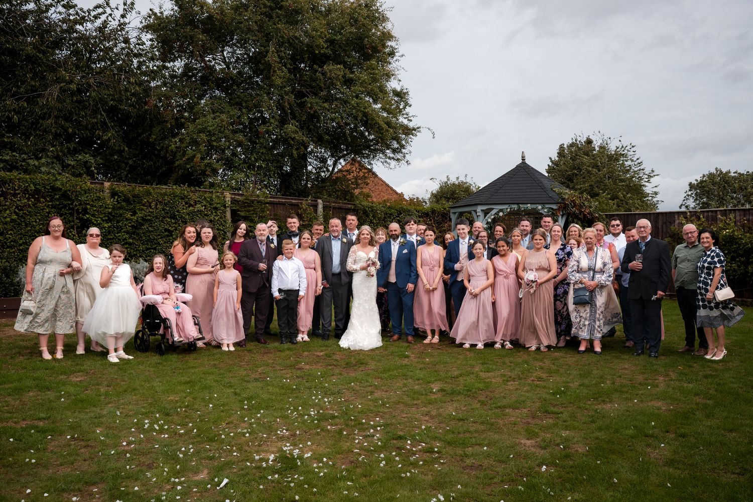 Large wedding group photo outdoors on green lawn with bride, groom, bridesmaids in pink, and guests near garden gazebo.