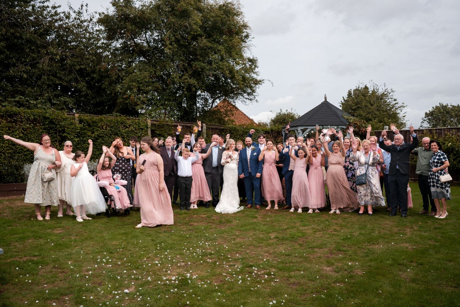 Large wedding party celebrates outdoors on green lawn, guests cheering with arms raised near a gazebo.