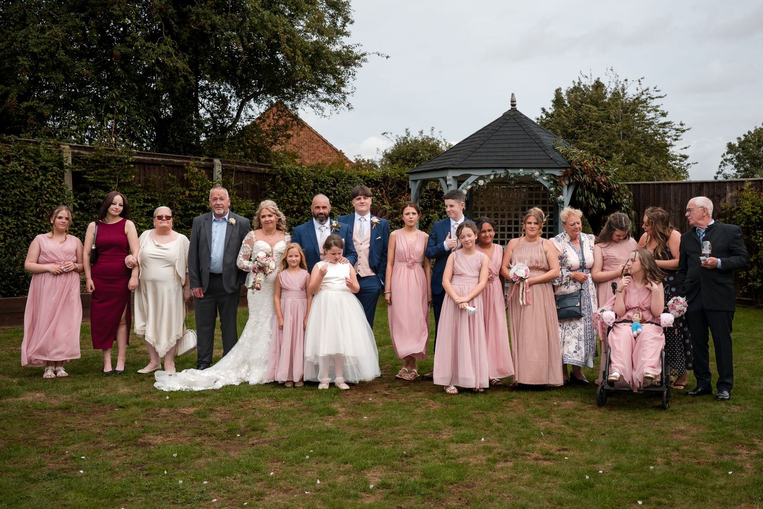 Wedding party posing outdoors in garden, bride in white gown with bridesmaids in pink dresses and groomsmen in suits.
