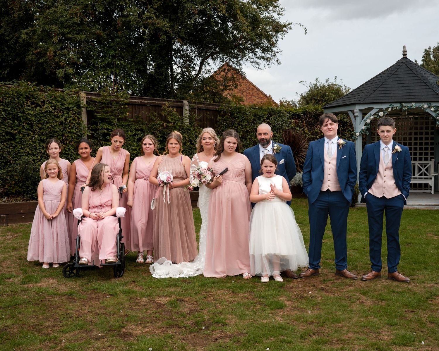 Wedding party poses outdoors, bride and groom with bridesmaids in pink dresses and groomsmen in navy suits on a garden lawn.