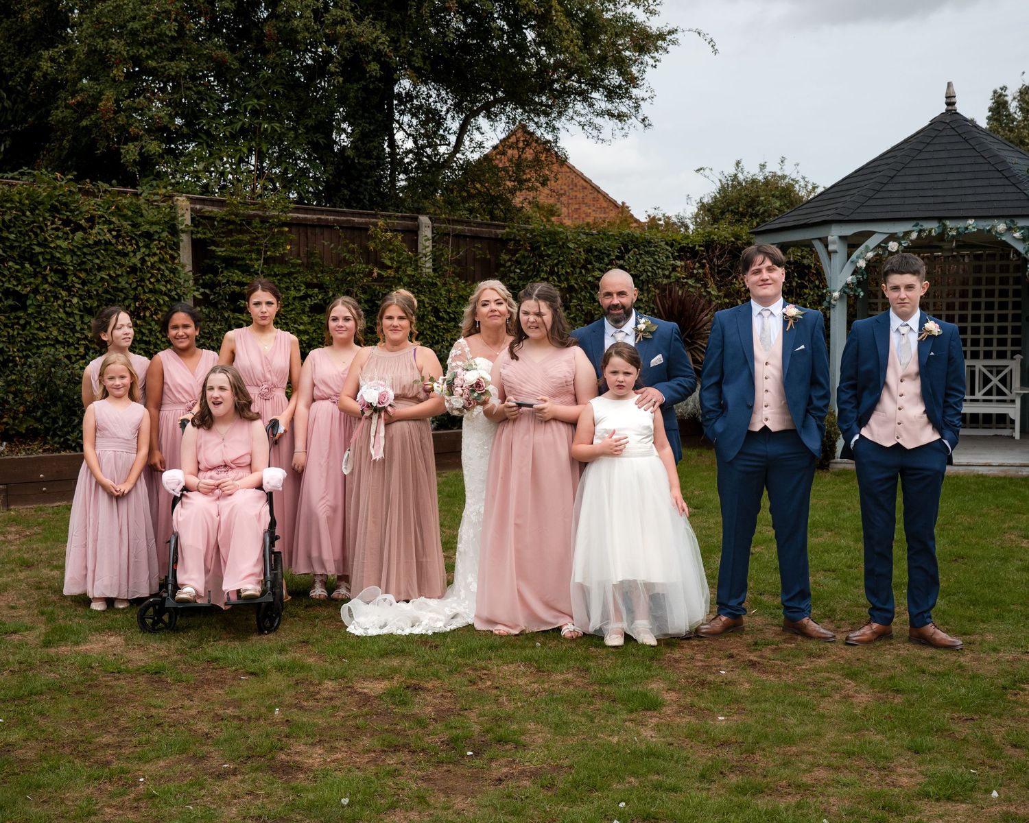Wedding party poses outdoors with bridesmaids in pink dresses and groomsmen in navy suits near a gazebo.
