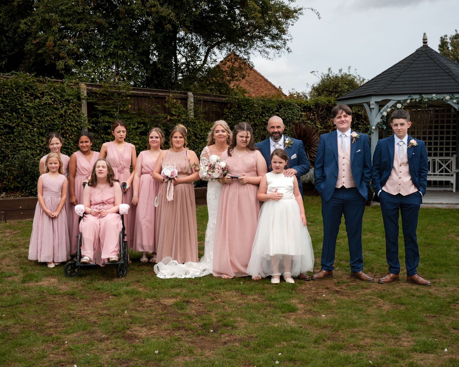Wedding group photo featuring bridesmaids in blush pink gowns and groomsmen in navy suits on a garden lawn.