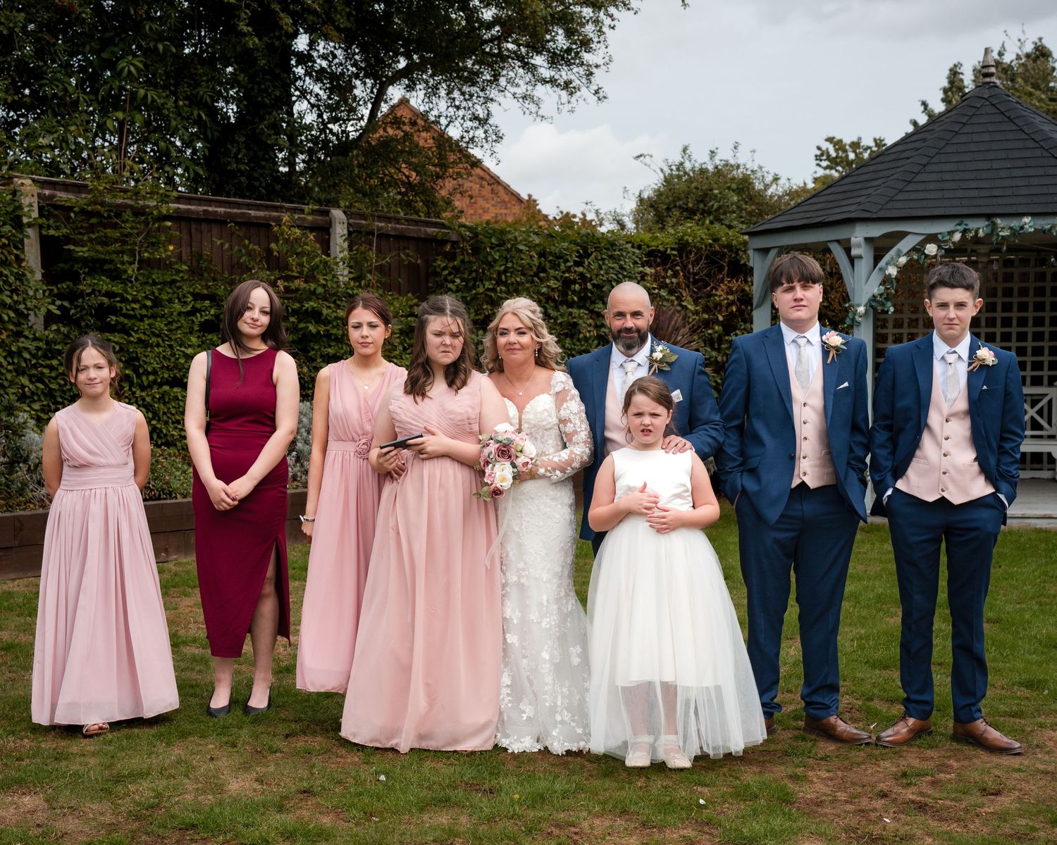 Wedding party posing outdoors, bride in white lace gown with bridesmaids in pink dresses and groomsmen in navy suits.