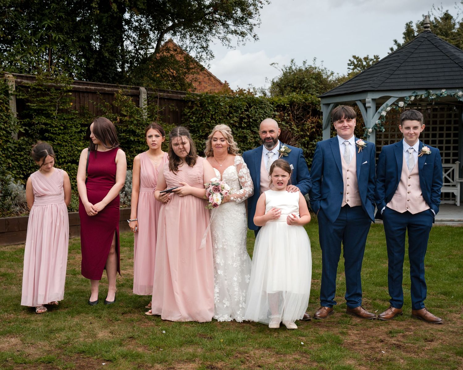Wedding party poses outdoors, bride in white gown with bridesmaids in pink dresses and groomsmen in navy suits by a gazebo.