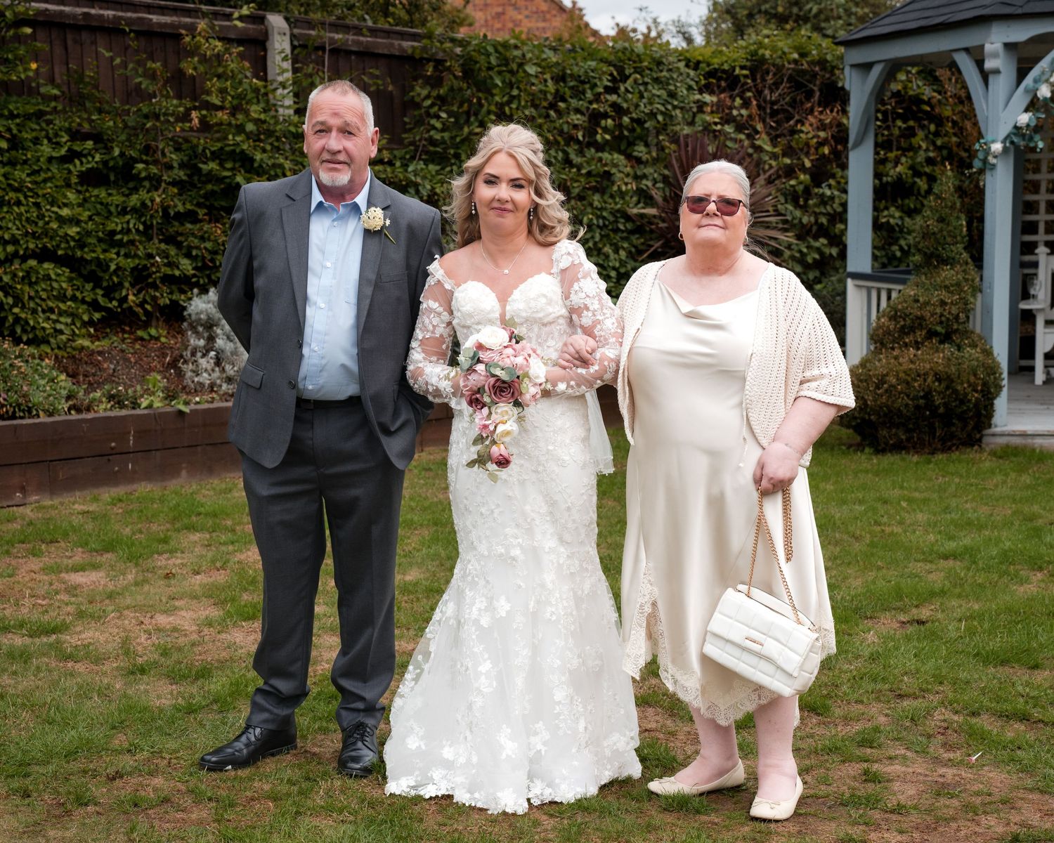 Bride in lace wedding dress holding bouquet poses with two guests in a garden wedding setting.