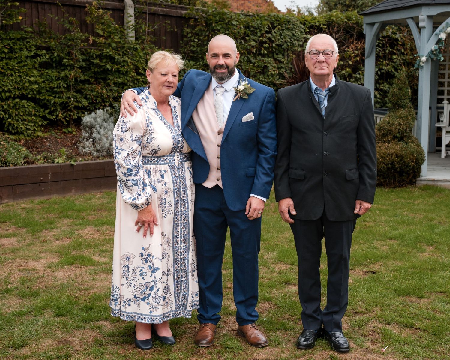 A groom in a blue suit poses with an older couple at an outdoor wedding reception in a garden setting.