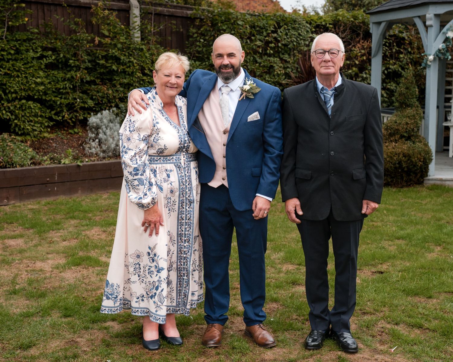 A groom in a blue suit poses with an older couple in a garden at a wedding celebration.