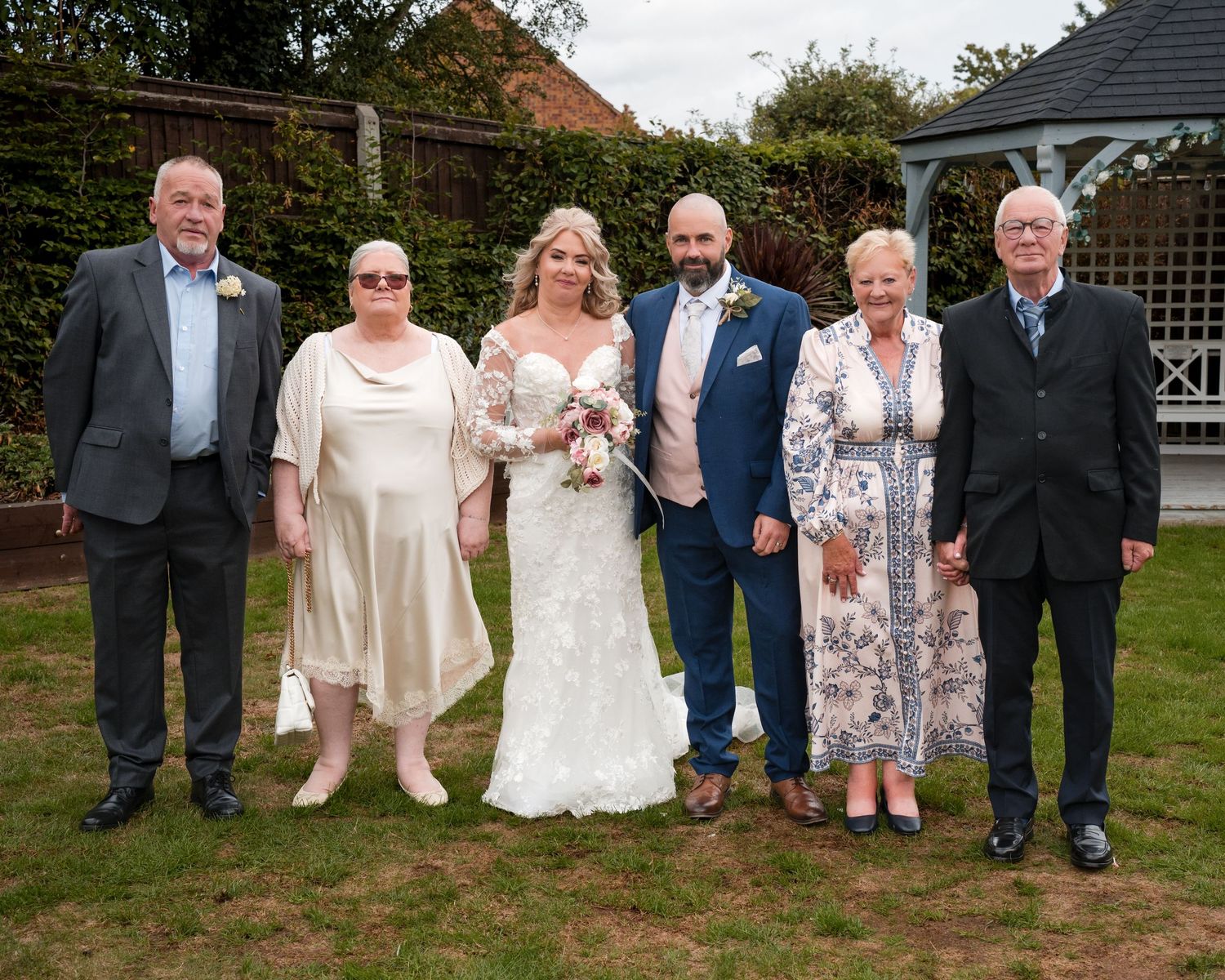 Wedding party of six posing outdoors in garden, bride in white lace gown with groom and family members dressed formally.