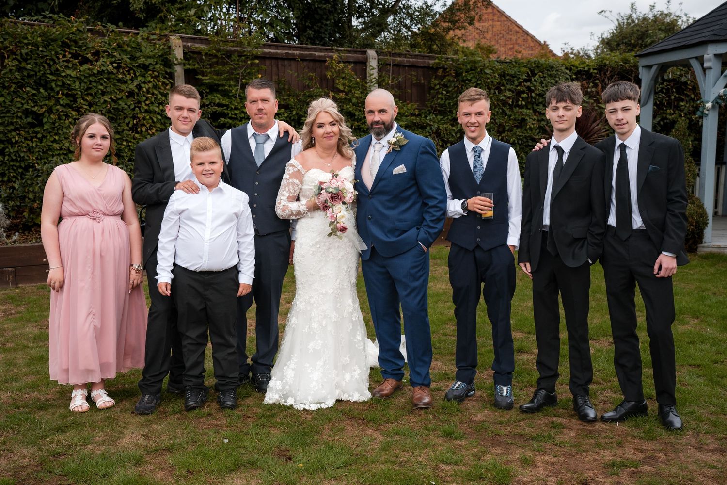 Wedding party posing outdoors on grass, bride in lace gown with groom in blue suit surrounded by family in formal attire.