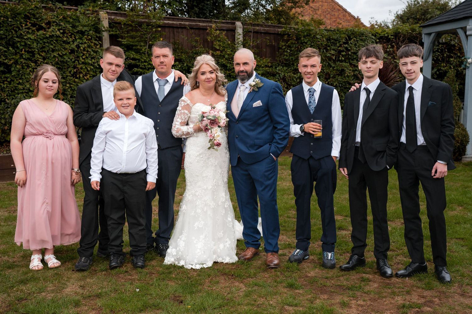Wedding party poses outdoors, bride in lace gown with groom in blue suit surrounded by family members in formal attire.
