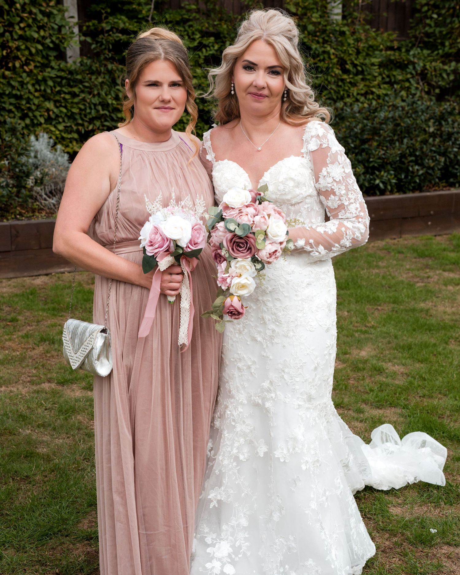Bride in white lace gown with bridesmaid in dusty pink dress holding floral bouquets outdoors at wedding.