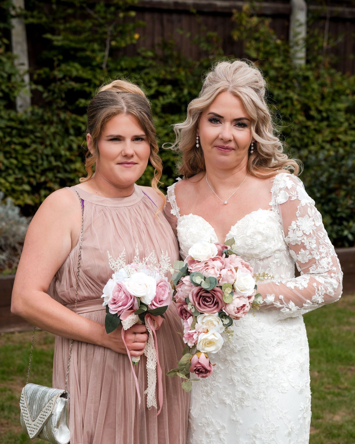 Bride in white lace gown holding pink bouquet poses with bridesmaid in dusty rose dress at outdoor wedding.