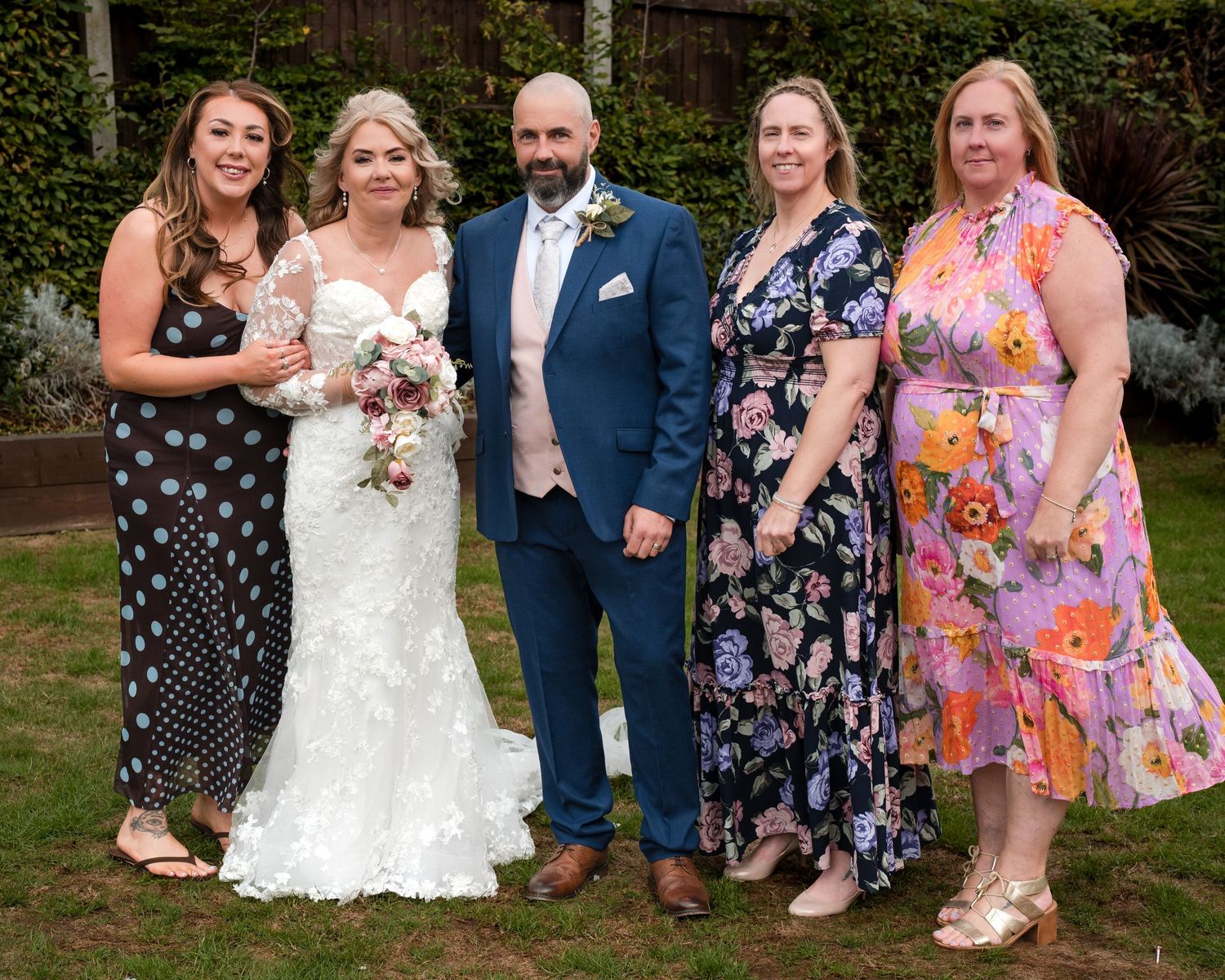 A bride and groom pose with three female guests in a garden setting during a wedding celebration.