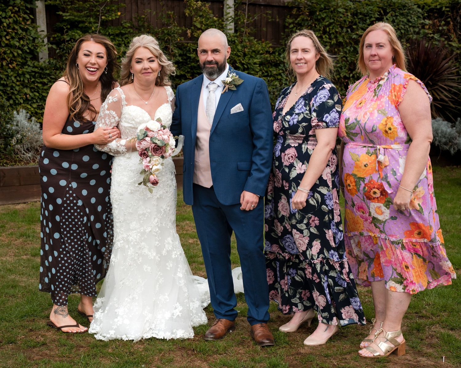 A bride and groom pose with three female guests in colorful dresses at an outdoor wedding celebration.