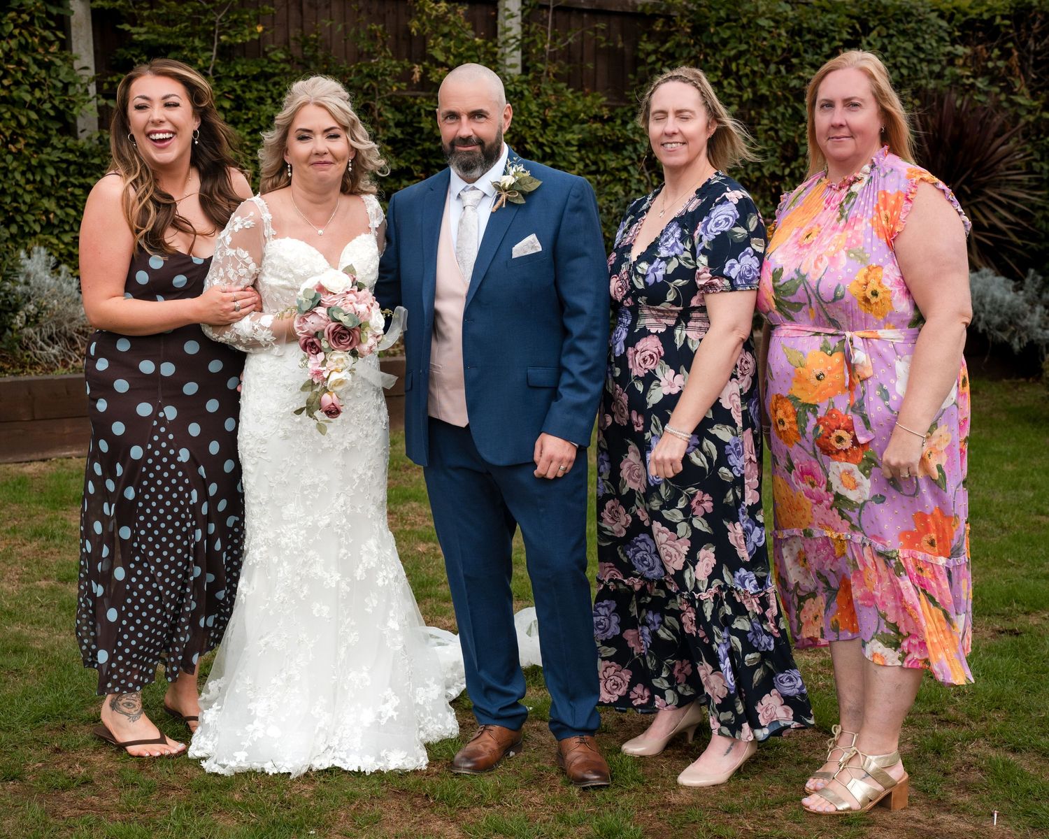A bride and groom pose with three female guests in a garden setting during an outdoor wedding celebration.