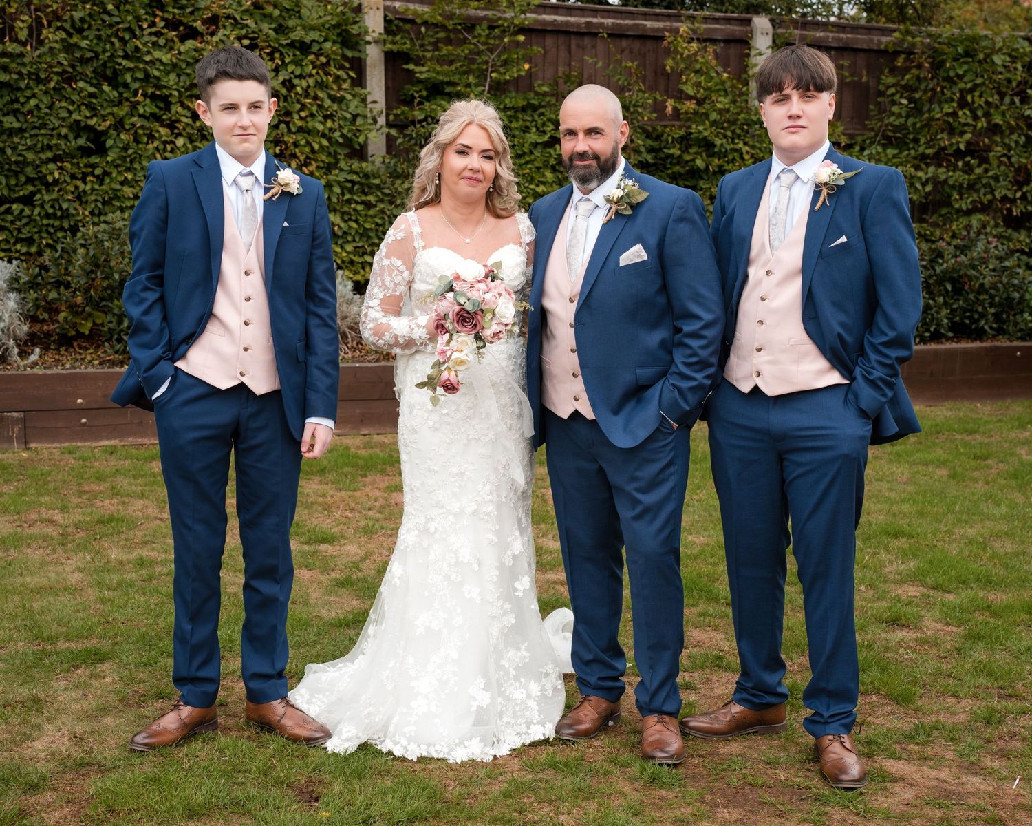 Wedding party posing outdoors, bride in lace gown with groom and two young men in navy blue suits with pink waistcoats.