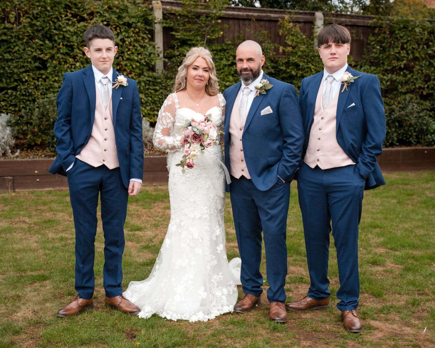 Bride in lace gown poses with groom and two young men in matching navy blue suits with blush waistcoats at wedding.