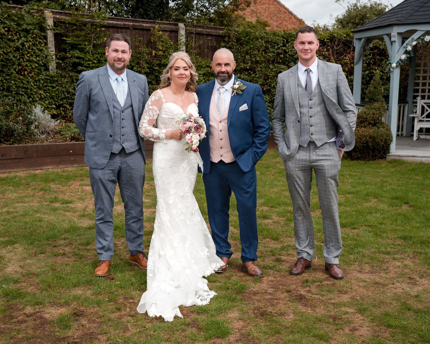 Bride in lace wedding dress poses with three men in suits on a garden lawn at an outdoor wedding ceremony.
