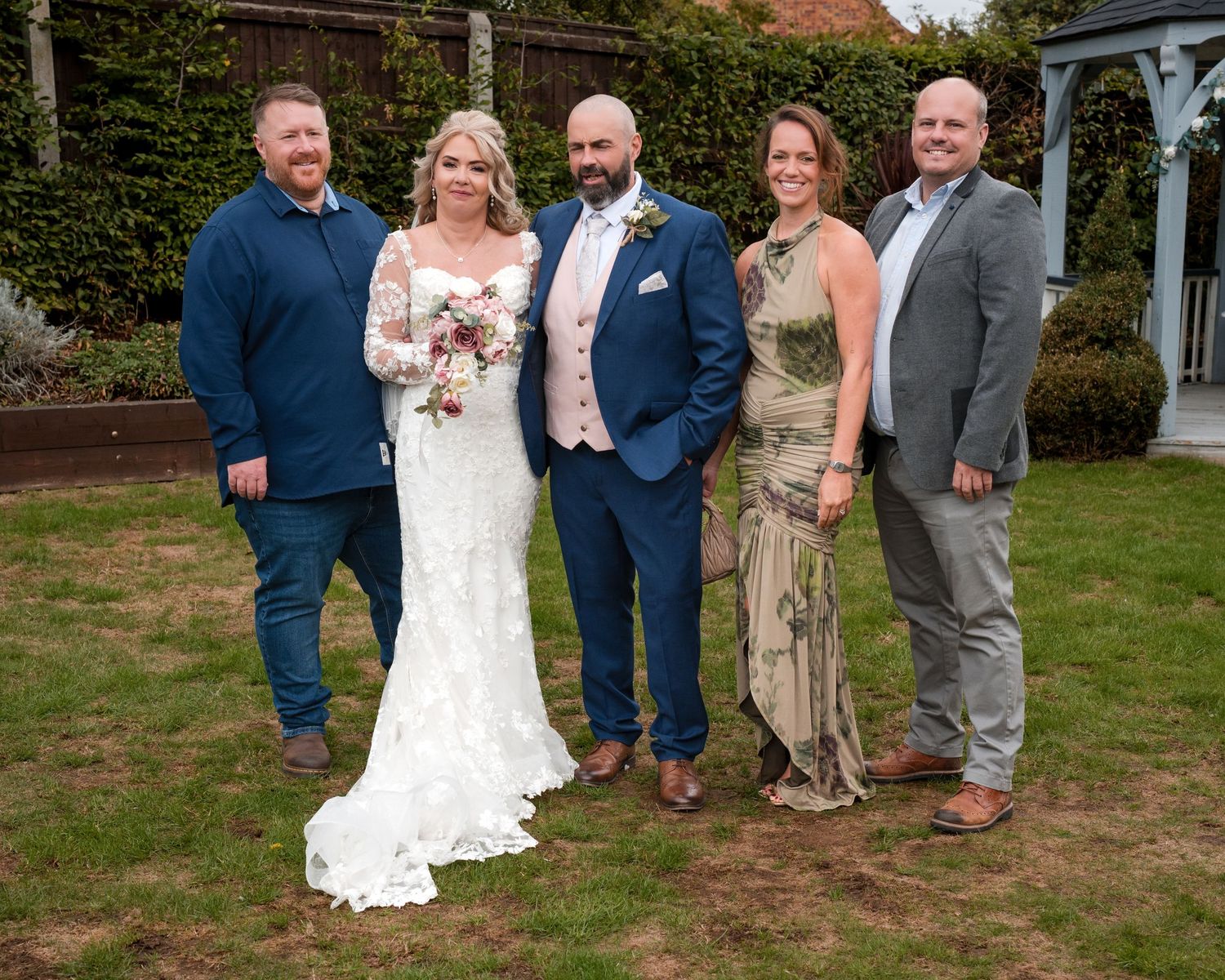 A bride and groom pose with three guests on a lawn at an outdoor wedding ceremony in a garden setting.