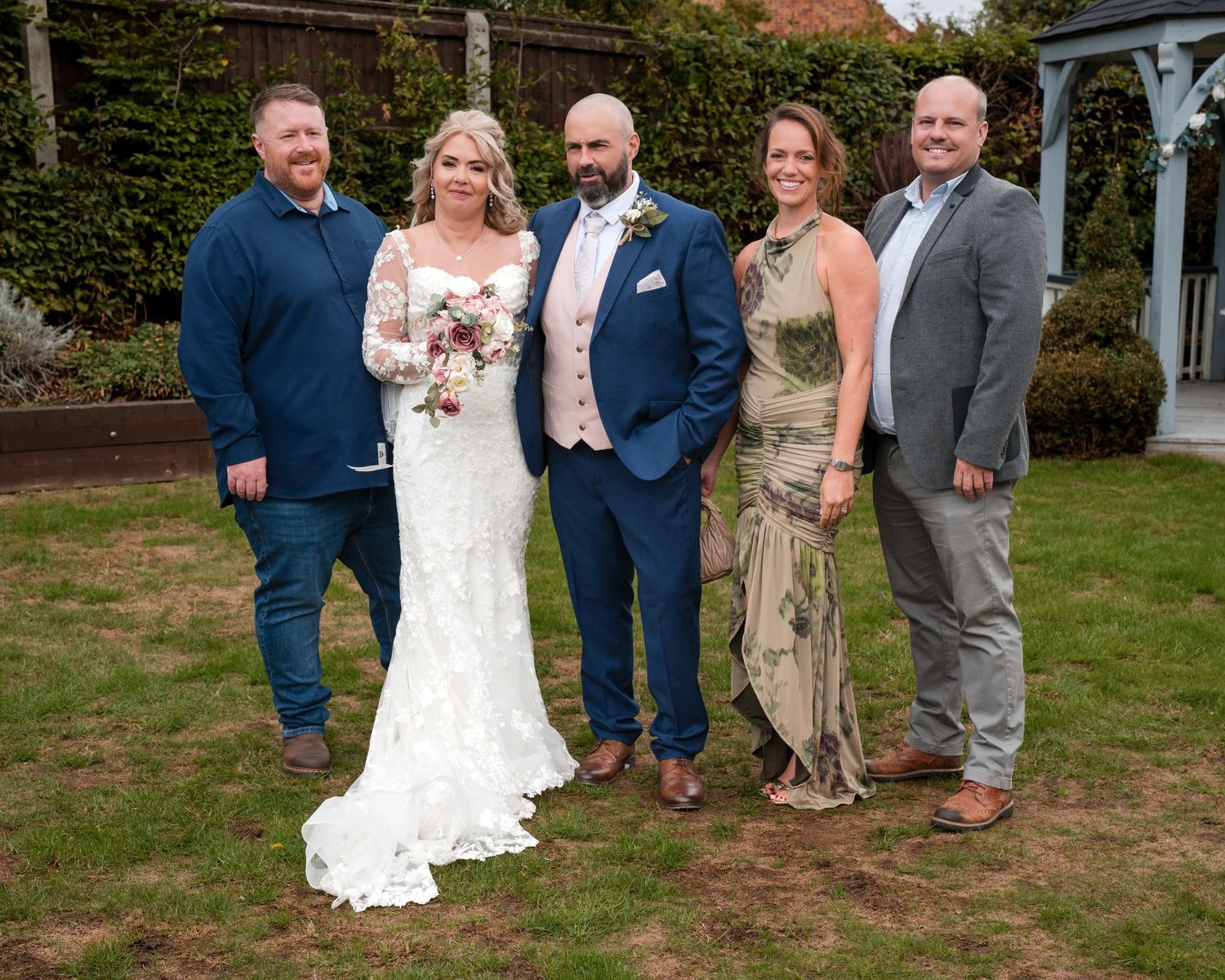 Wedding couple in white dress and blue suit pose with three guests outdoors at a garden venue.