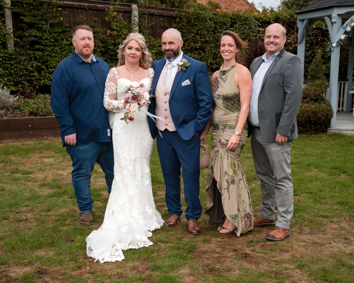 Bride and groom in blue suit pose with three guests on green lawn at outdoor wedding venue.