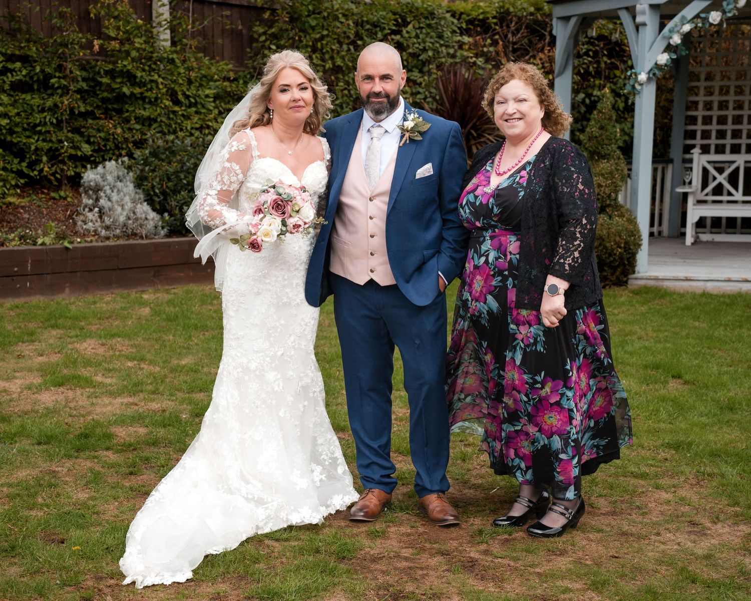Bride in lace gown and groom in blue suit pose with a guest in floral dress at an outdoor wedding ceremony.