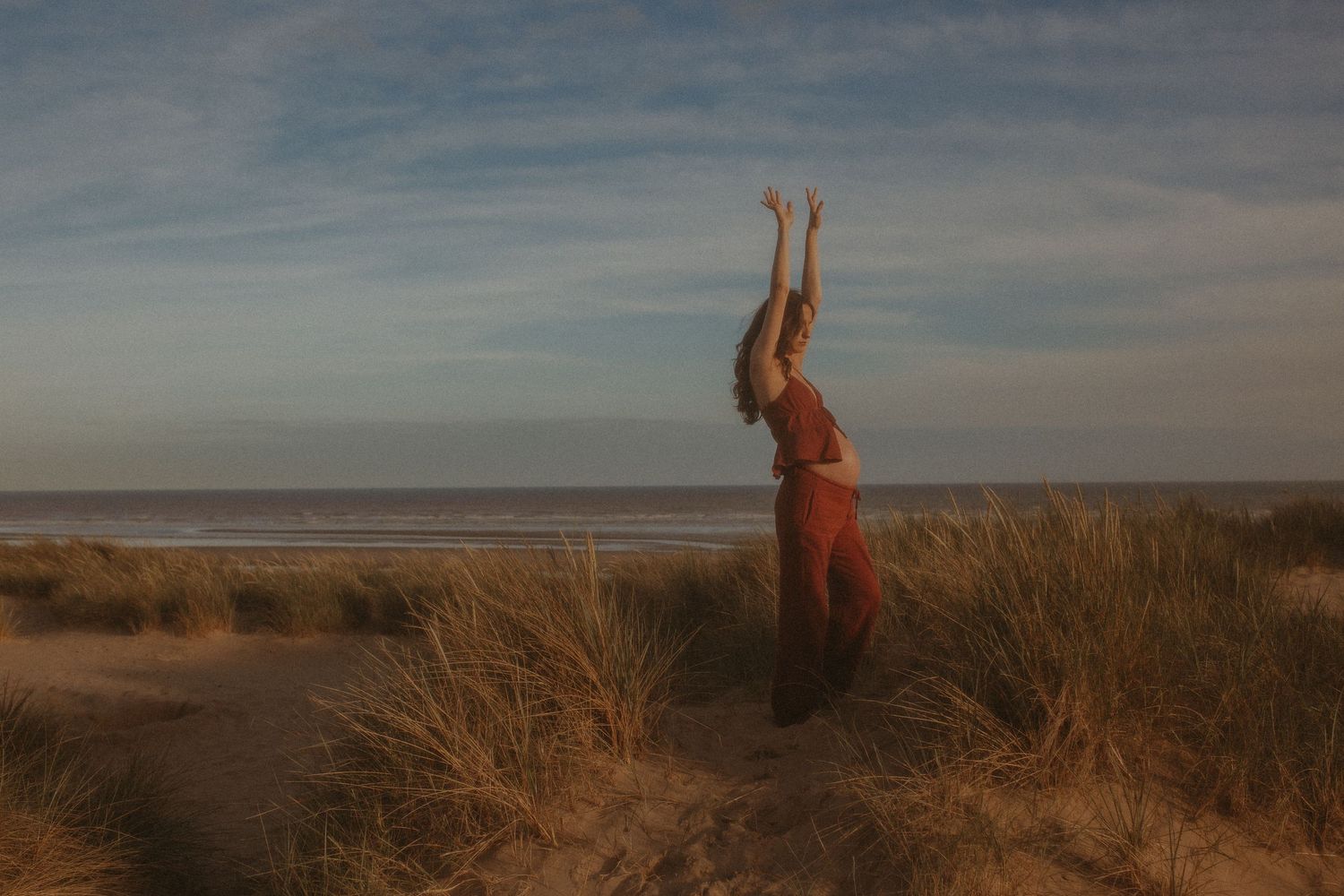 Woman in red dress raising arms on sandy beach dunes with ocean horizon and blue sky backdrop.