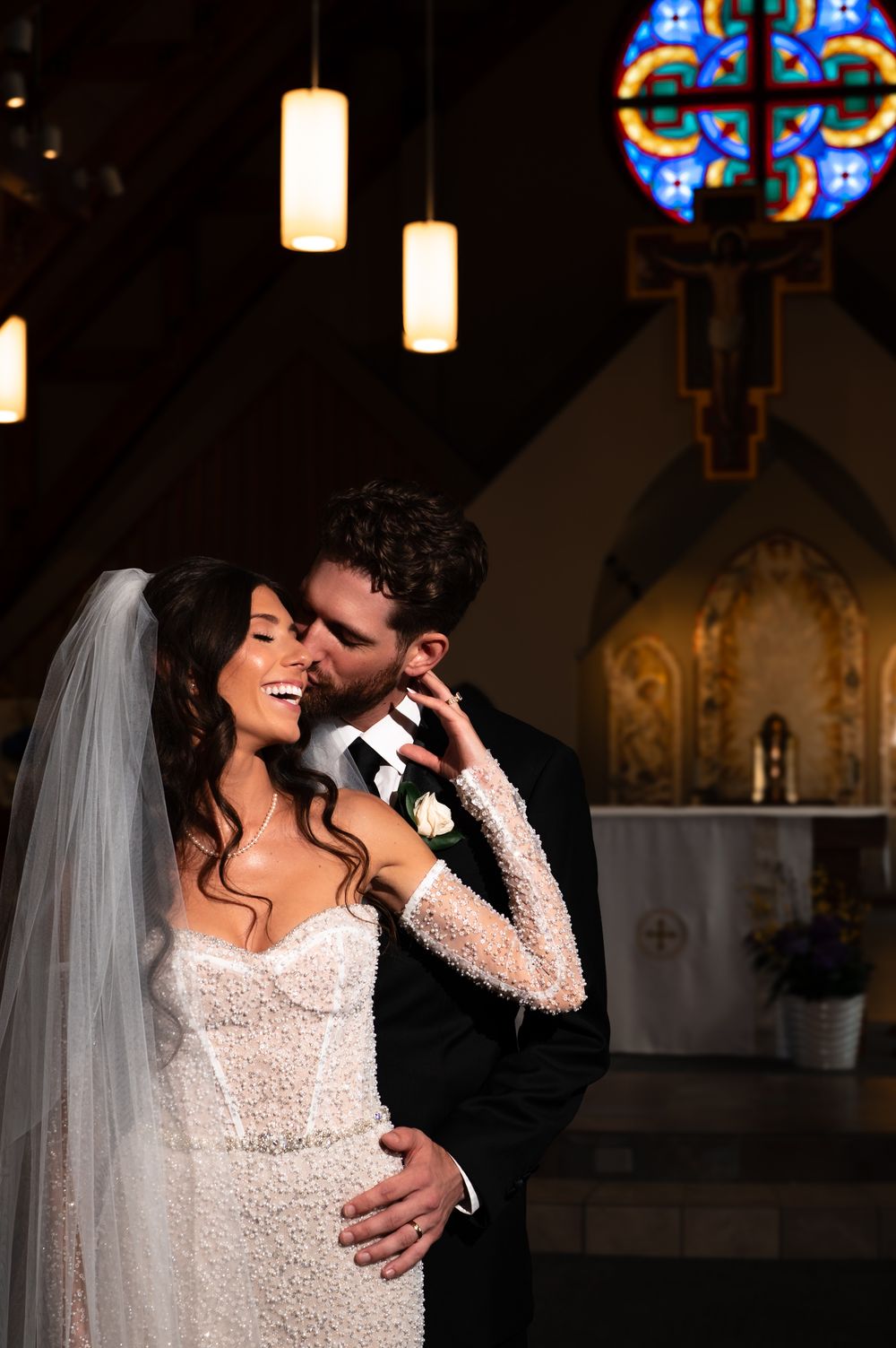 Bride and groom share romantic moment in church, bride in lace gown with veil, groom in black suit, stained glass window behind.