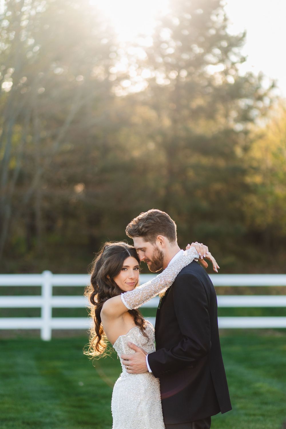 Bride and groom embrace outdoors at golden hour, white fence and trees in background, wedding portrait.