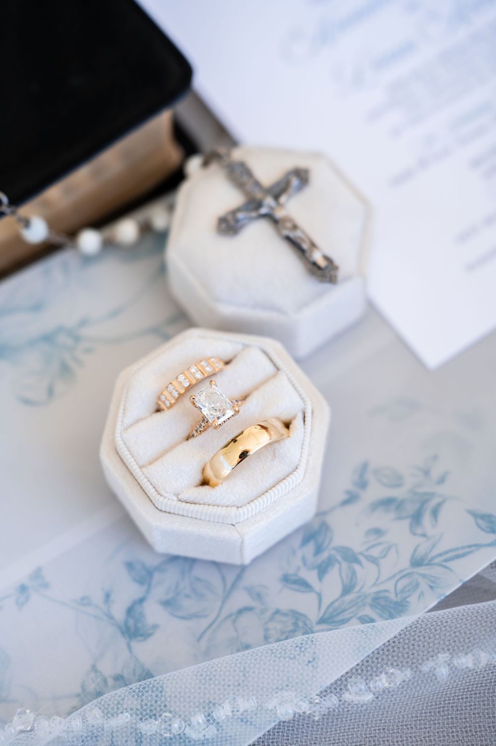 Gold wedding bands and diamond engagement ring displayed in a white velvet ring box beside a silver crucifix cross.