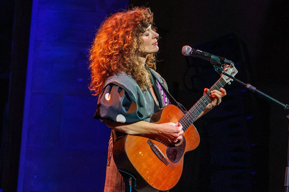 Red-haired female musician playing acoustic guitar and singing into microphone on stage during live performance.