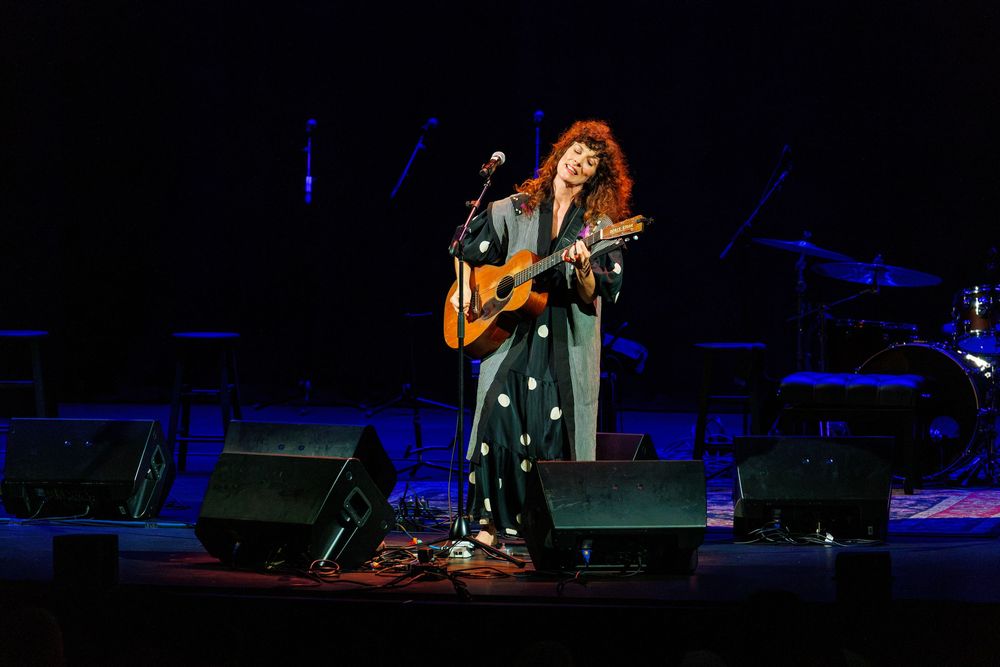 Female musician performing on stage with acoustic guitar, wearing a floral dress under concert lights.