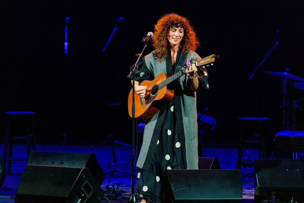 A curly red-haired woman plays acoustic guitar on stage, wearing a dark kimono with polka dots, performing at a concert.