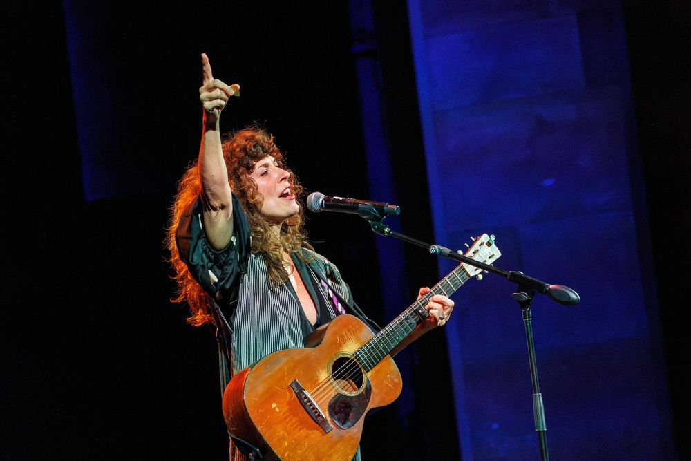 Female musician with curly red hair plays acoustic guitar on stage, pointing upward at a live concert performance.