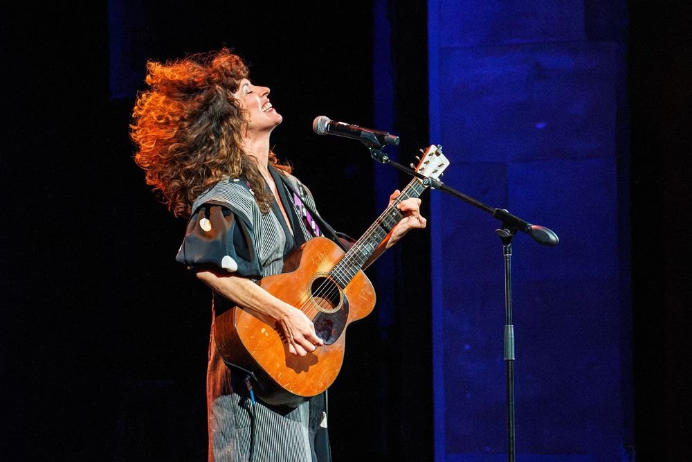 A curly-haired woman plays acoustic guitar and sings into a microphone on a dimly lit stage.