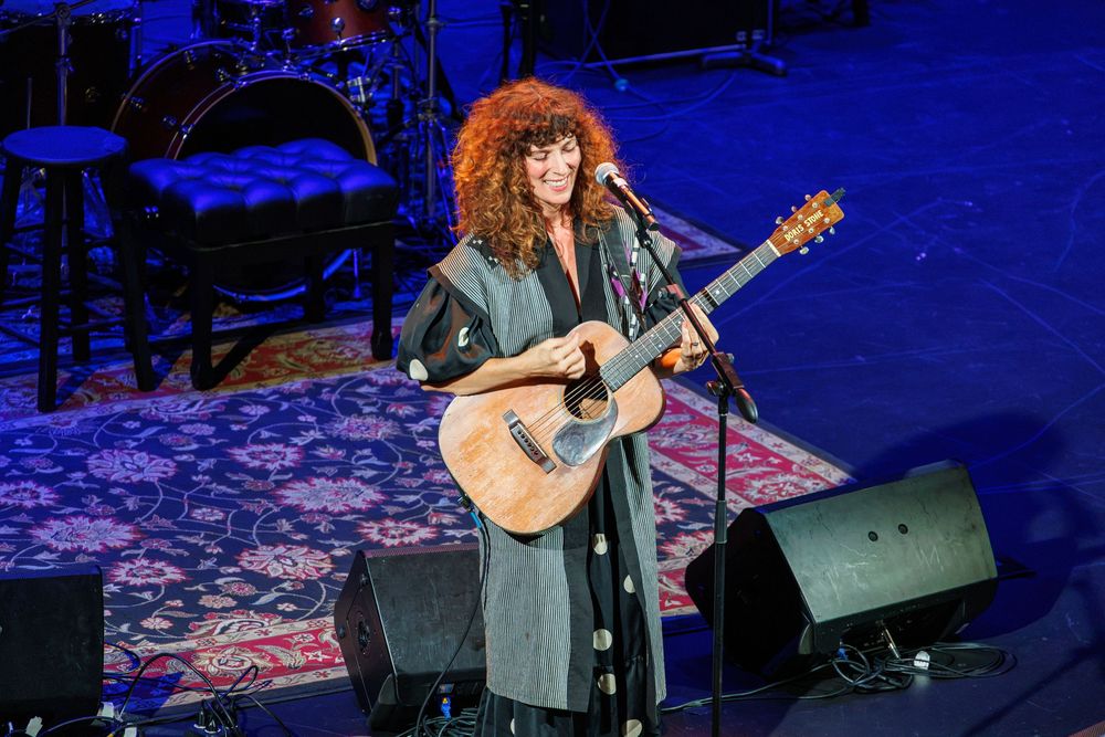 A curly-haired woman performs on stage playing an acoustic guitar while wearing a striped outfit under concert lights.