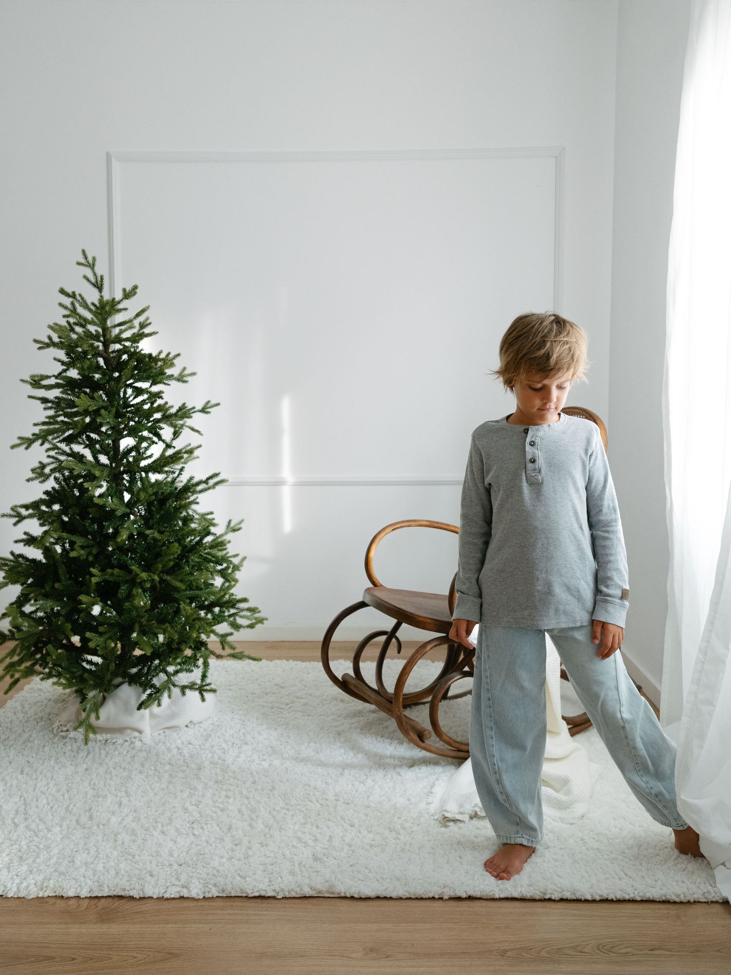 A young boy in gray pajamas stands barefoot on a white rug beside a decorated Christmas tree in a bright minimalist room.