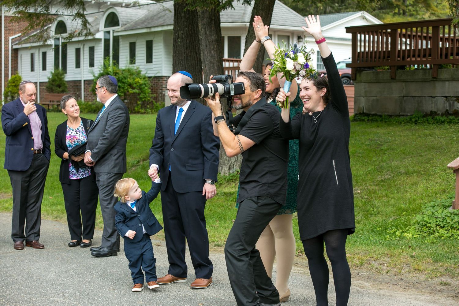 Wedding photographer capturing bride raising bouquet while guests watch outdoor ceremony.