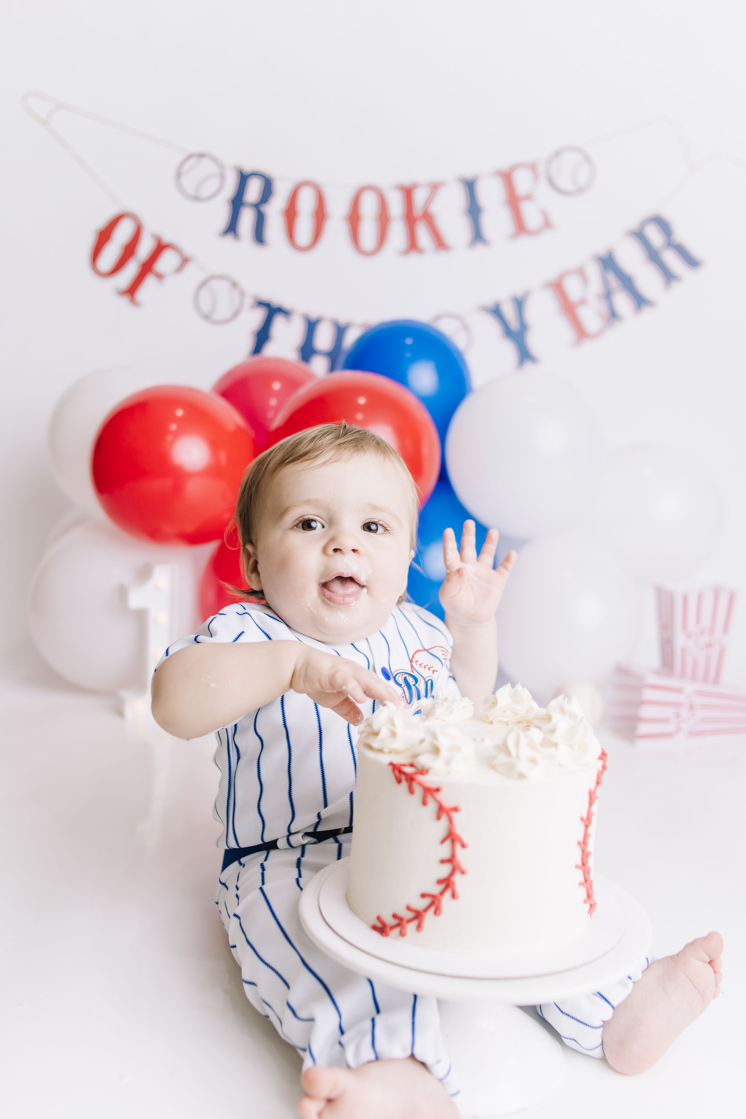 Baby in baseball outfit celebrates first birthday with baseball cake under Rookie of the Year banner and colorful balloons.
