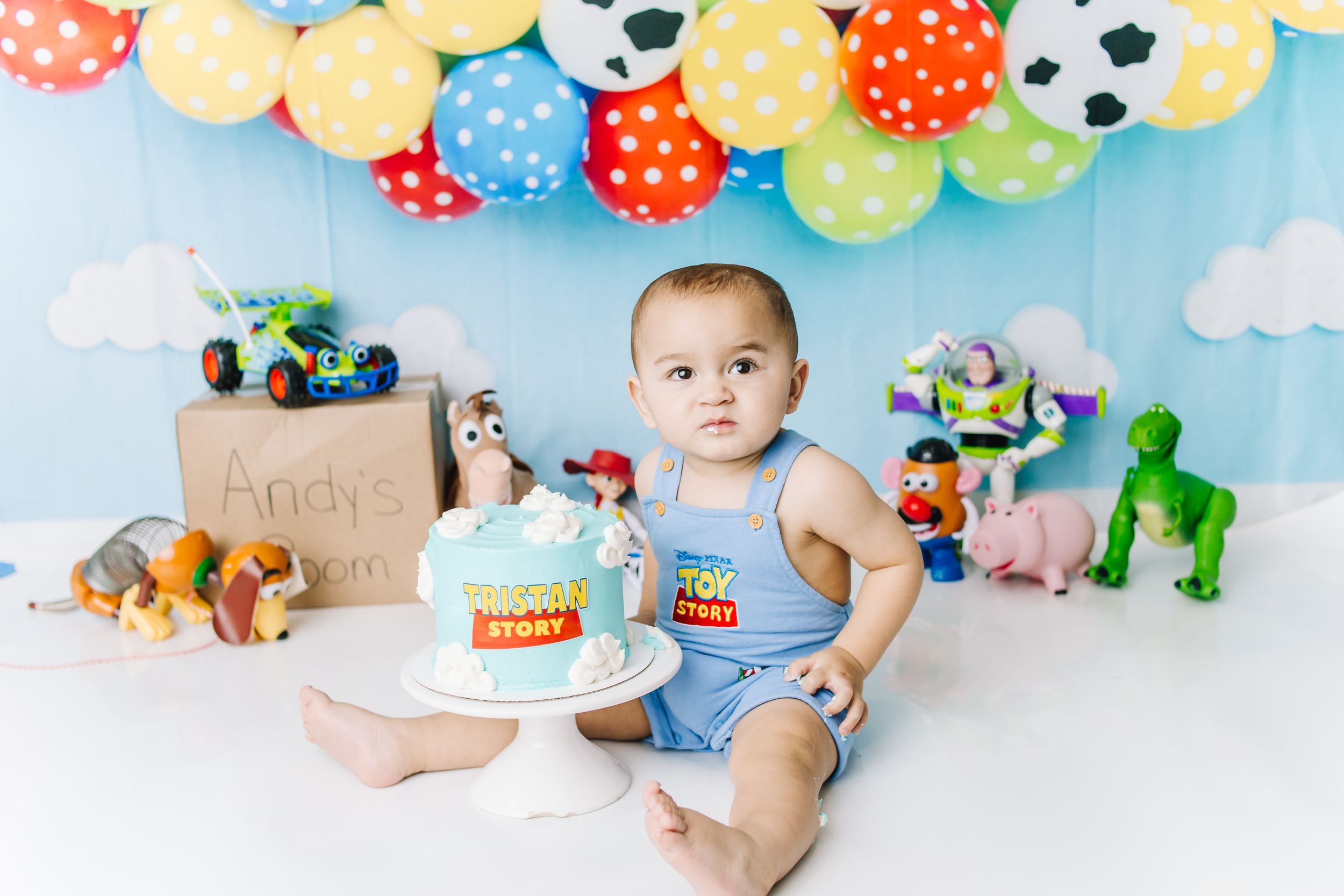 Baby boy in blue romper sits with Toy Story themed first birthday cake surrounded by colorful balloons and character toys.