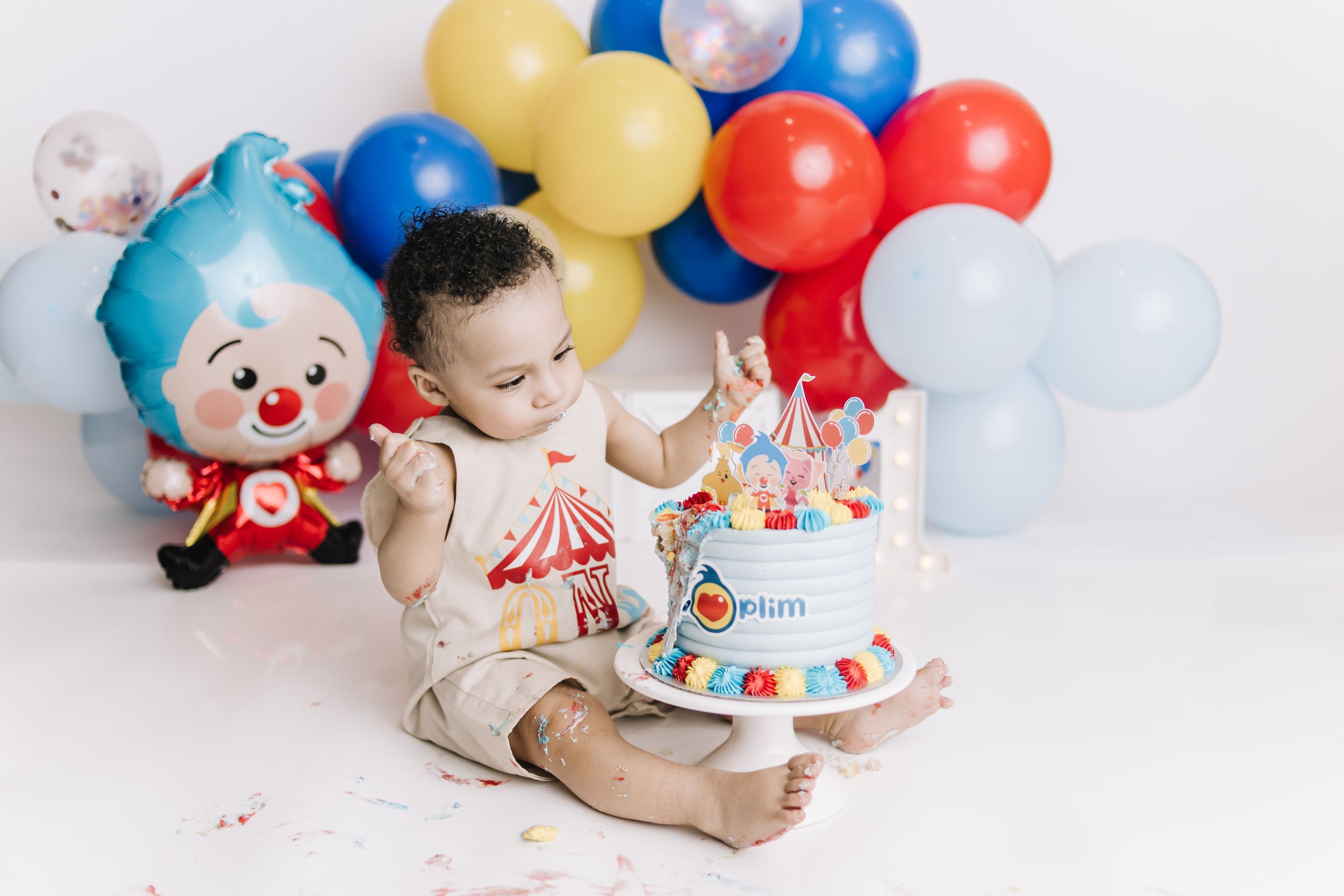 Baby boy celebrating first birthday with colorful circus cake, clown toy, and balloons in festive smash cake photoshoot.