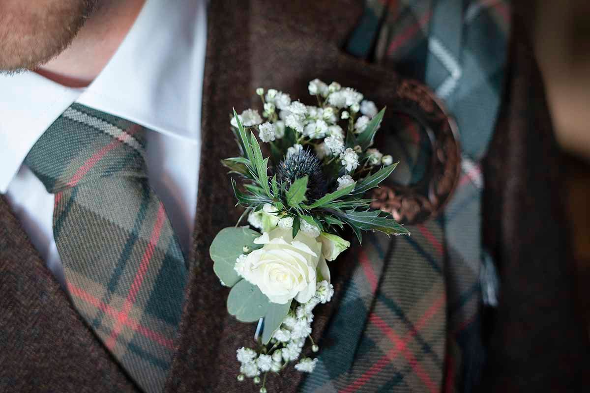 Close-up of a white rose and thistle boutonniere on a tartan plaid suit lapel at a Scottish wedding.