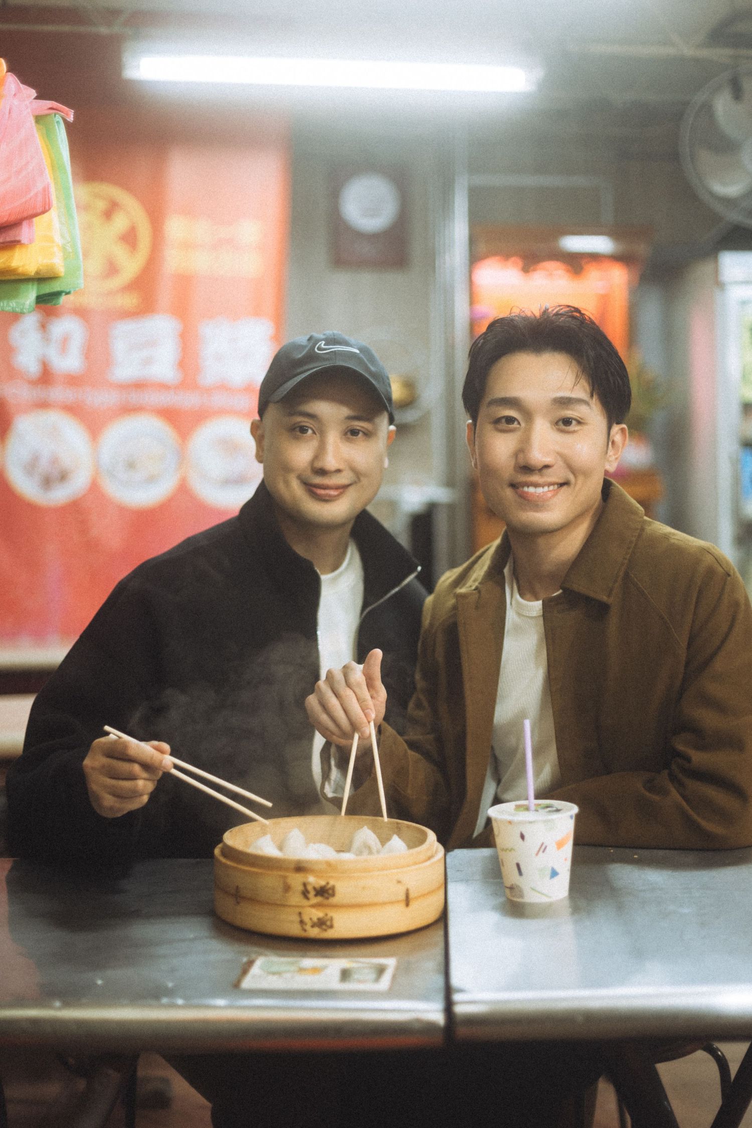 Two young men smile at camera while enjoying dim sum dumplings with chopsticks at a cozy Asian restaurant.