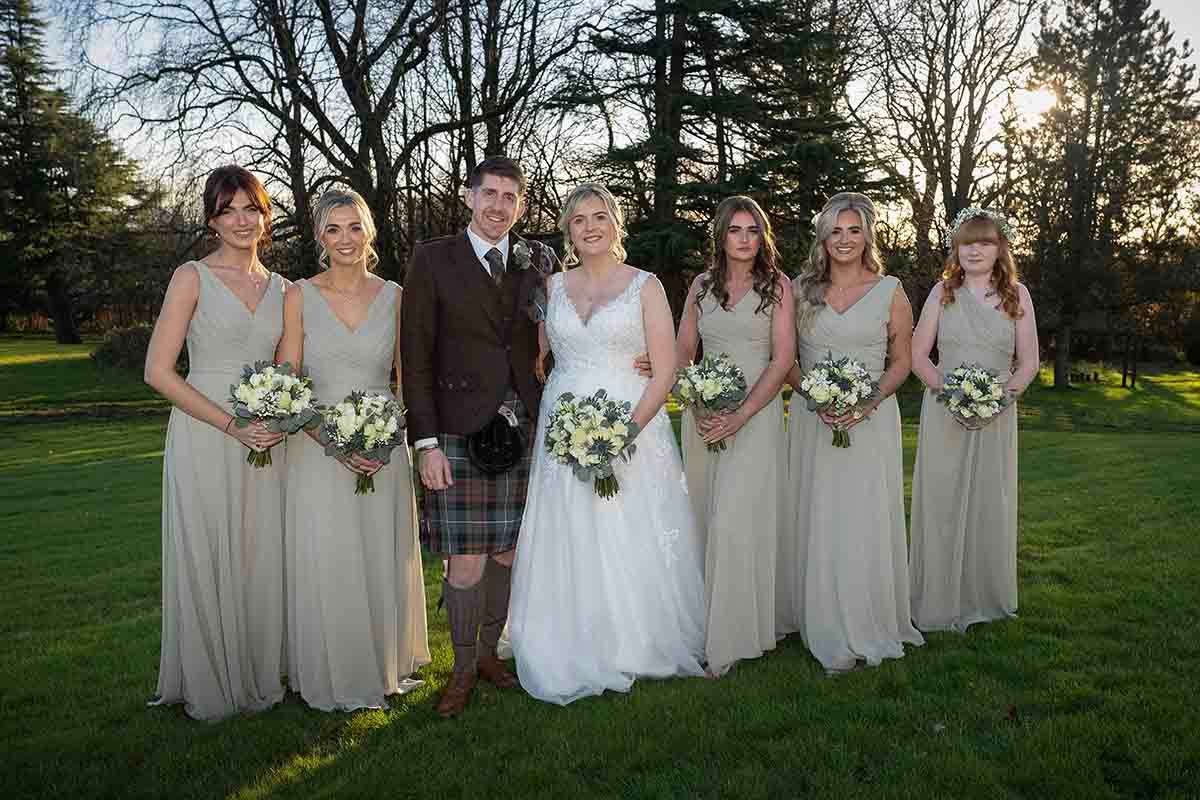 Groom in tartan kilt with bride in white gown and five bridesmaids in champagne dresses holding white bouquets outdoors at Hetland Hall