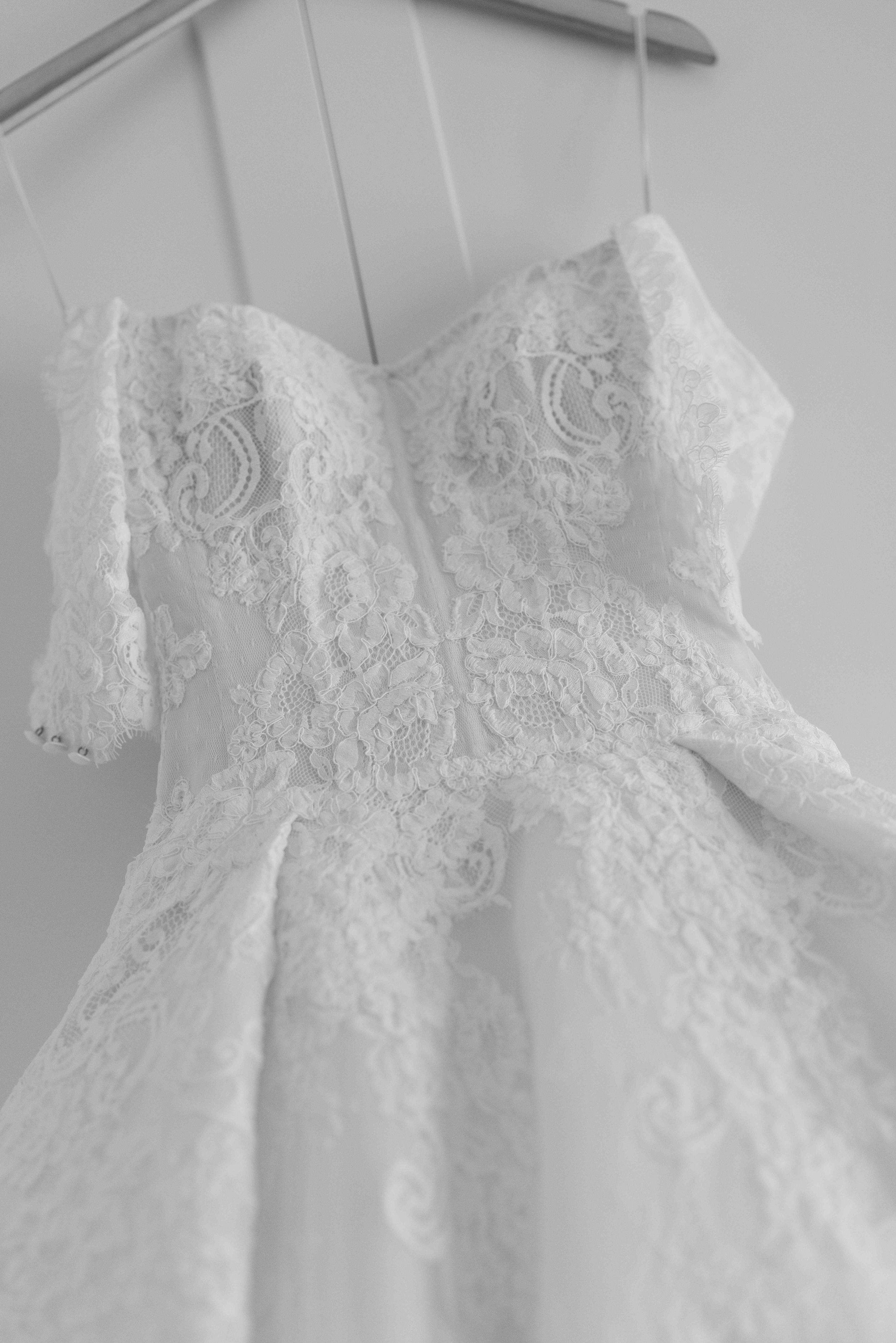Black and white photo of an elegant off-shoulder lace wedding dress hanging on a wooden hanger.
